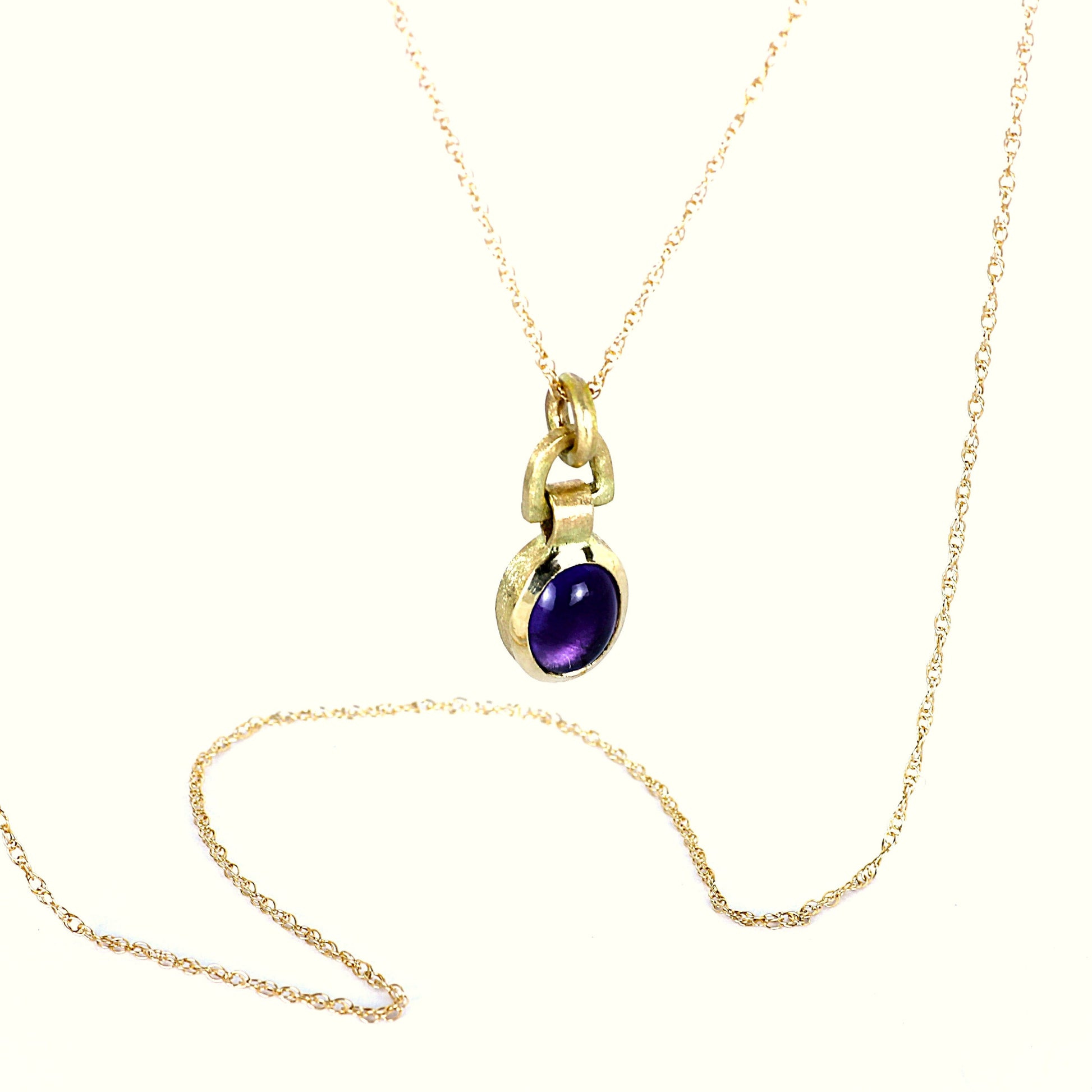 A gold chain necklace with a small gold pendant featuring a round purple gemstone, displayed on a clean white background.