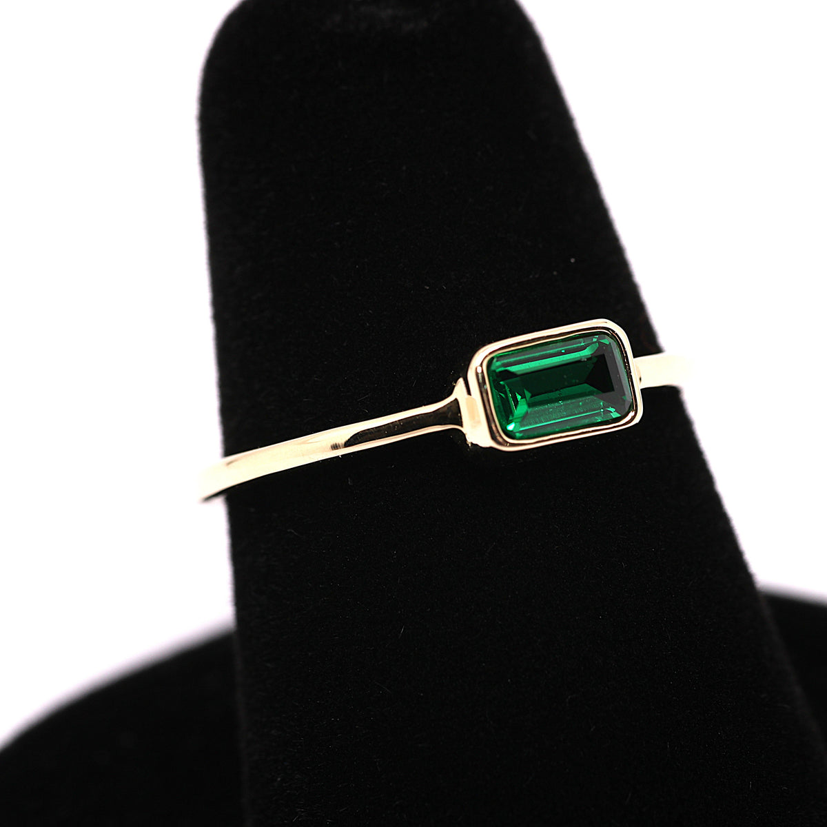 A gold ring with a rectangular green emerald-cut gemstone, displayed on a black velvet ring stand against a white background.